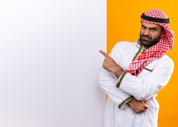 Homme d'affaires arabe en vêtements traditionnels debout près de panneau d'affichage vierge pointant avec le doigt dessus avec un visage en colère sur un mur orange