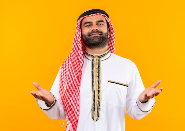 Homme d'affaires arabe en vêtements traditionnels avec sourire confiant en levant les bras debout sur un mur orange