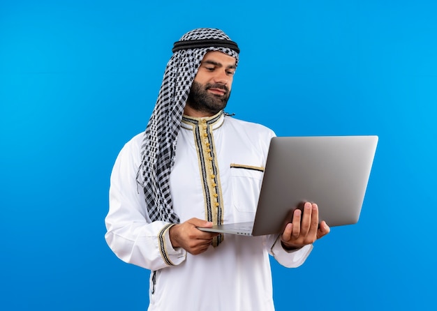 Homme d'affaires arabe en vêtements traditionnels tenant un ordinateur portable en le regardant avec le sourire sur le visage debout sur un mur bleu