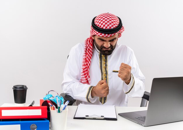 Homme d'affaires arabe en vêtements traditionnels travaillant avec un ordinateur portable serrant le poing avec une expression agressive mécontent et frustré assis à la table au bureau