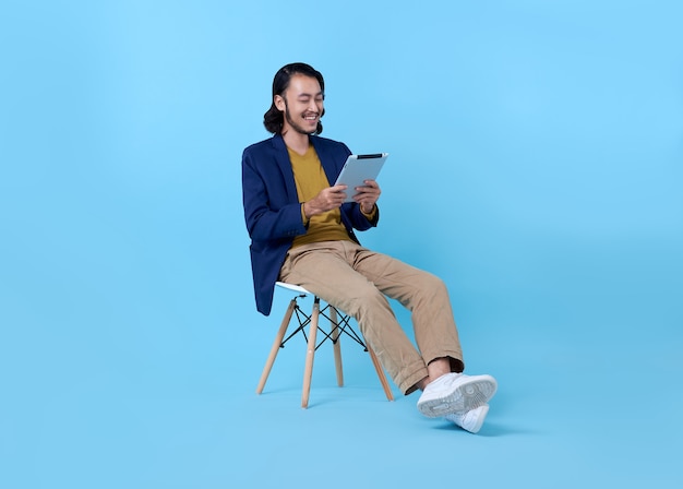 Homme d'affaires asiatique heureux souriant à l'aide d'une tablette numérique alors qu'il était assis sur une chaise bleu vif.