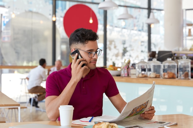 Un homme d'affaires attrayant et prospère écrit le dernier numéro de journal, se concentre sur une publication intéressante pendant la pause-café, discute de quelque chose avec son partenaire via un téléphone portable, écrit dans un cahier