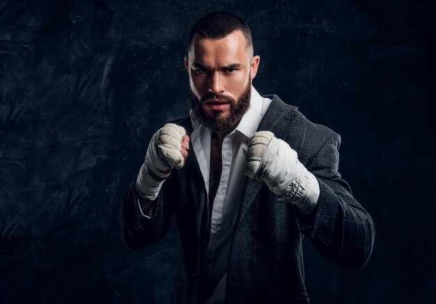 Un homme d'affaires barbu en colère en costume et des gants de kickboxing protecteurs pose pour le photographe dans un studio photo sombre.