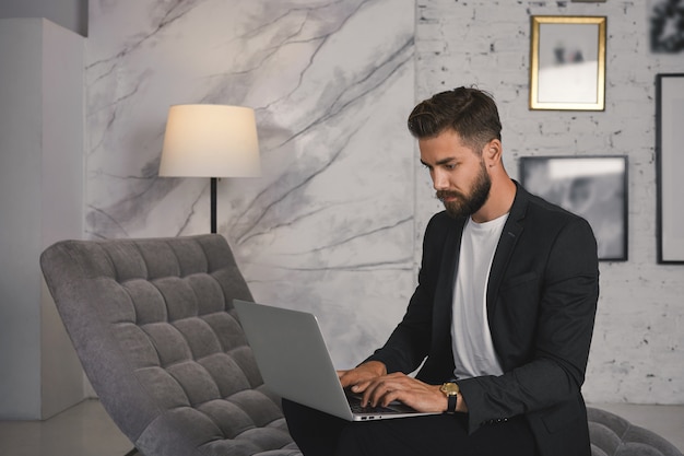 Homme d'affaires barbu élégant assis sur un canapé confortable avec un ordinateur portable sur son ordinateur portable, en tapant une lettre commerciale par e-mail, après avoir concentré son regard sérieux. Technologie, communication et affaires