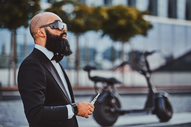 Un homme d'affaires barbu intelligent se tient près de son vélo électrique tout en fumant un vaporisateur.