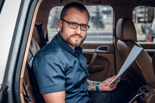 Un homme d'affaires barbu portant des lunettes avec un tatouage sur le bras est assis sur le siège arrière d'une voiture.