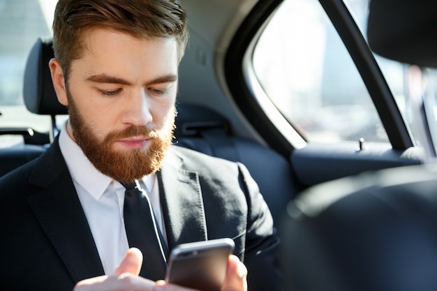 Homme d'affaires barbu sérieux en costume regardant un téléphone portable dans sa main