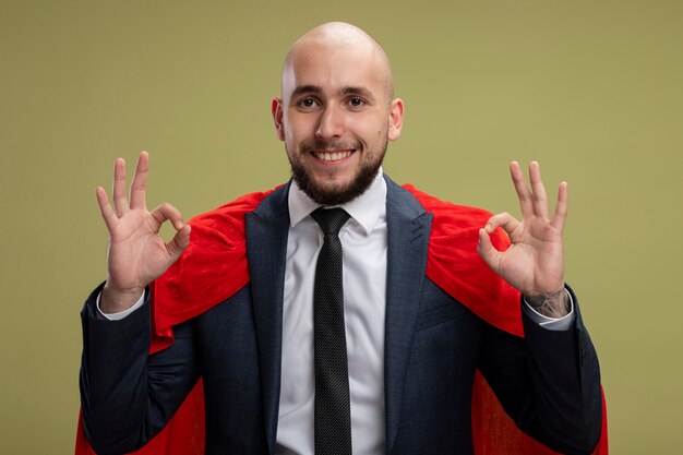 Homme d'affaires barbu super héros en cape rouge à la recherche de sourire joyeusement montrant signe ok debout sur un mur vert clair