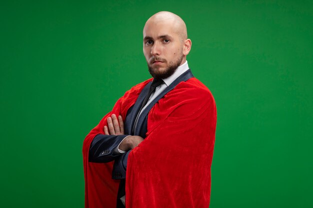 Homme d'affaires barbu super héros en cape rouge avec un visage sérieux avec les bras croisés sur la poitrine debout sur le mur vert