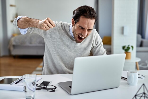 Homme d'affaires en colère criant de frustration et sur le point de frapper son ordinateur portable après avoir lu de mauvaises nouvelles tout en travaillant à la maison