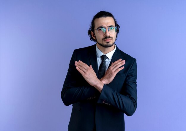 Homme d'affaires en costume noir et lunettes à l'avant faisant panneau d'arrêt croisant les mains debout sur le mur bleu