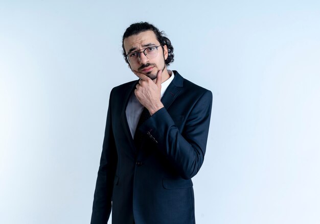 Homme d'affaires en costume noir et lunettes à l'avant avec la main sur le menton pensant debout sur un mur blanc