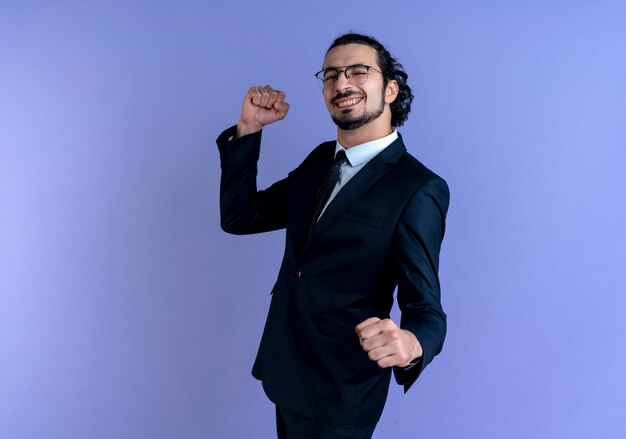 Homme d'affaires en costume noir et lunettes à l'avant serrant les poings heureux et excité debout sur le mur bleu