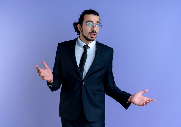 Homme d'affaires en costume noir et lunettes à la confusion et incertitude des bras sur les côtés debout sur le mur bleu