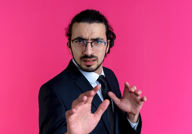 Homme d'affaires en costume noir et lunettes faisant un geste de défense avec les mains avec une expression dégoûtée debout sur un mur rose