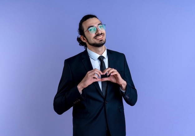 Homme d'affaires en costume noir et lunettes faisant le geste du cœur avec les doigts sur sa poitrine à l'avant avec le sourire debout sur le mur bleu