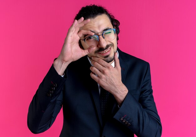 Homme d'affaires en costume noir et lunettes faisant signe ok à l'avant avec le sourire à travers ce panneau debout sur un mur rose