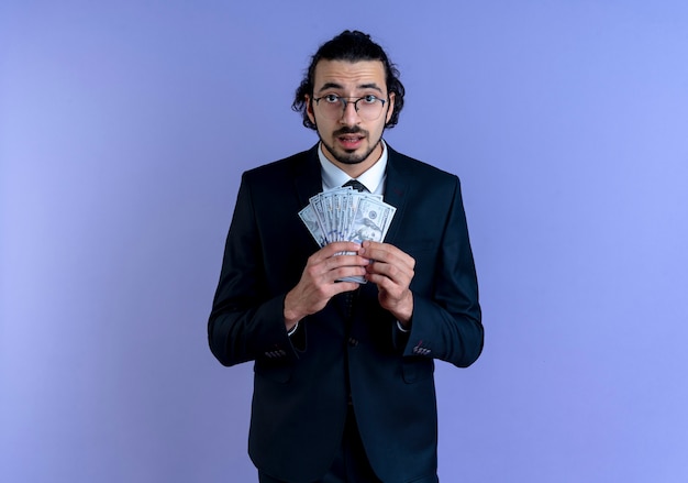 Homme d'affaires en costume noir et lunettes montrant de l'argent à l'avant confus debout sur un mur bleu