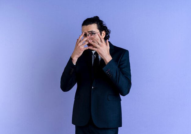 Homme d'affaires en costume noir et lunettes peur couvrant les yeux avec les doigts regardant à travers les doigts debout sur le mur bleu