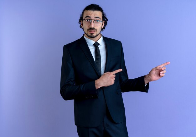 Homme d'affaires en costume noir et lunettes pointant avec l'index sur le côté à la confusion debout sur le mur bleu