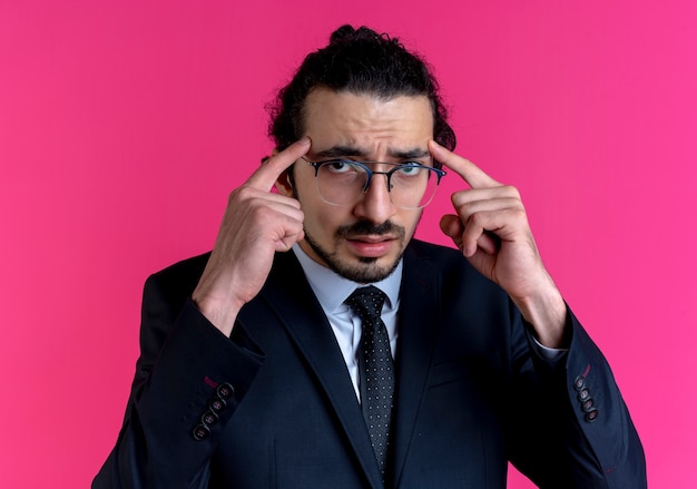 Homme d'affaires en costume noir et lunettes pointant ses tempes à la confusion debout sur le mur rose