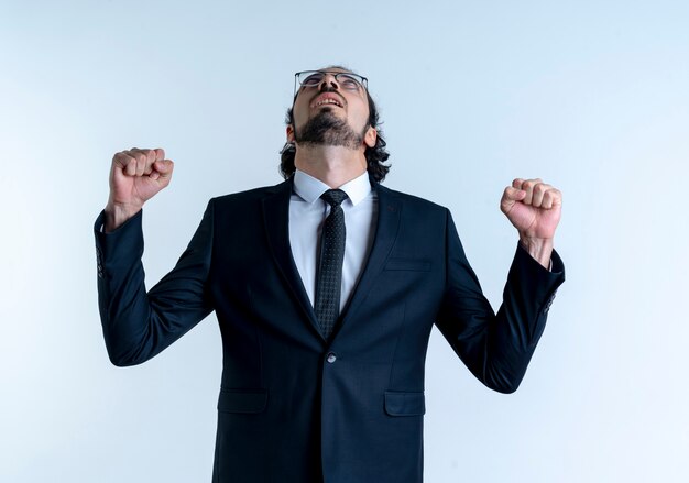 Homme d'affaires en costume noir et lunettes à la recherche de poings serrés heureux et excité debout sur un mur blanc