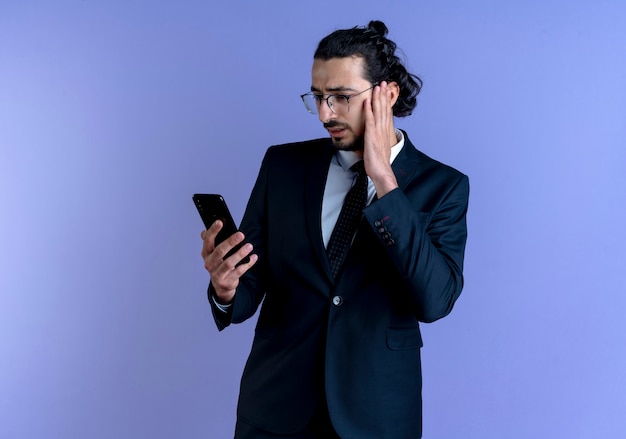 Homme d'affaires en costume noir et lunettes regardant l'écran de son smartphone avec une expression de confusion debout sur un mur bleu