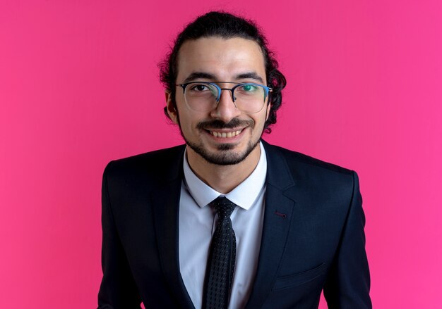 Homme d'affaires en costume noir et lunettes regardant vers l'avant souriant avec visage heureux debout sur un mur rose