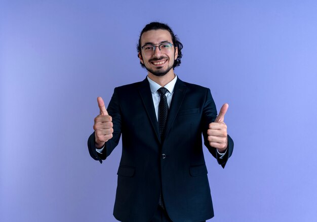 Homme d'affaires en costume noir et lunettes souriant à l'avant montrant les pouces vers le haut debout sur le mur bleu