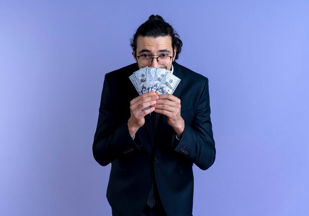 Homme d'affaires en costume noir et lunettes tenant de l'argent à la recherche de surprise et étonné debout sur le mur bleu