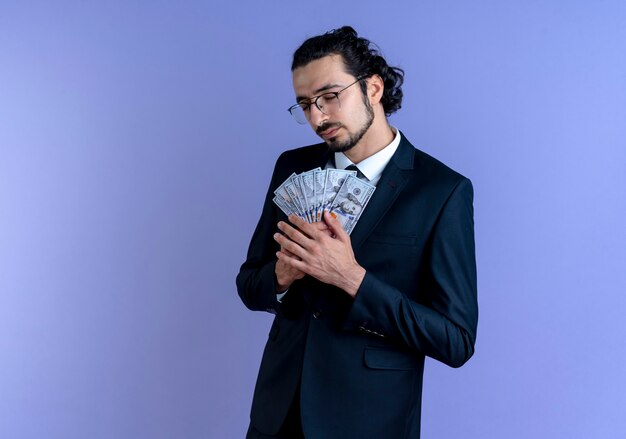 Homme d'affaires en costume noir et lunettes tenant de l'argent en se sentant reconnaissant avec les yeux fermés debout sur le mur bleu