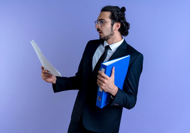 Homme d'affaires en costume noir et lunettes tenant le dossier regardant des documents avec un visage sérieux debout sur un mur bleu