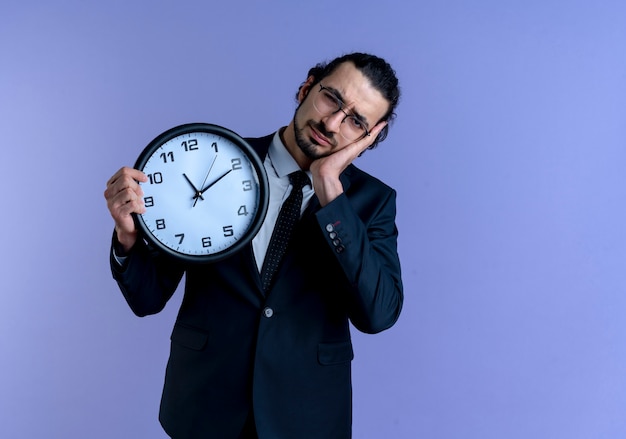 Homme D'affaires En Costume Noir Et Lunettes Tenant Horloge Murale à La Fatigue Et Surmené Faisant Le Geste De Sommeil Debout Sur Le Mur Bleu