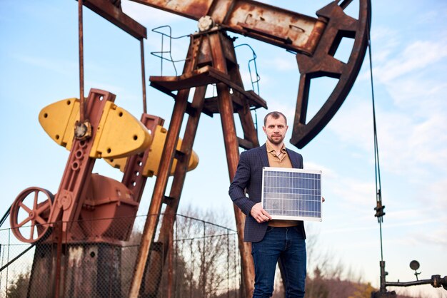 Homme d'affaires debout sur un champ pétrolifère tenant un mini module solaire à côté d'une plate-forme pétrolière