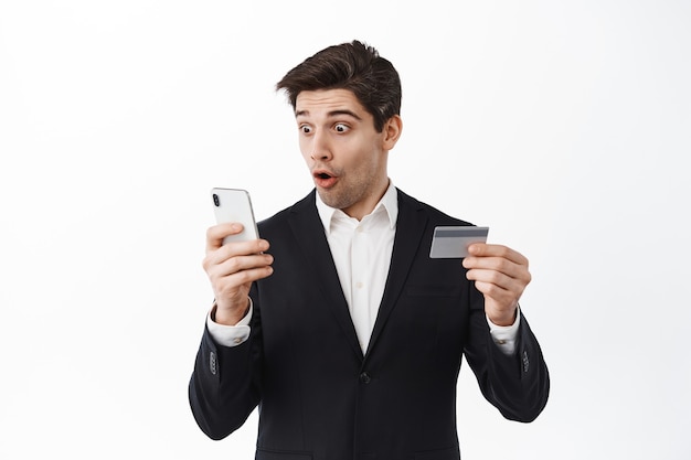 Un homme d'affaires enthousiaste en costume regarde son téléphone, lit un écran avec un visage étonné et tient une carte de crédit, se connecte à l'application de banque en ligne, debout sur un mur blanc