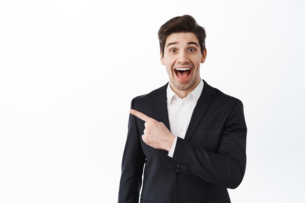 Homme d'affaires excité, entrepreneur d'entreprise en costume haletant étonné, pointant de côté l'espace de copie avec un visage souriant impressionné, vérifiant l'affaire géniale, mur blanc