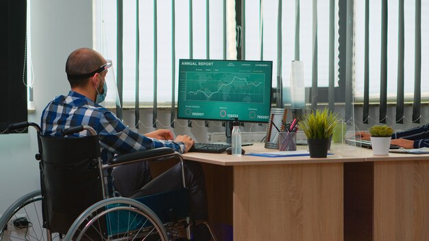 Homme d'affaires handicapé assis en fauteuil roulant avec masque de protection nettoyant les mains avant de vérifier les données financières dans le bureau moderne de l'entreprise. Indépendant handicapé avec visière respectant la distance sociale