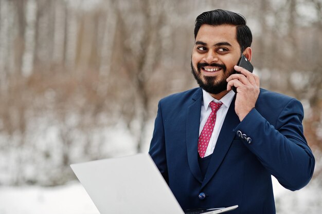 Homme d'affaires indien élégant barbe en costume posé à la journée d'hiver en plein air avec un ordinateur portable aux mains parlant au téléphone mobile