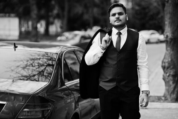 Homme d'affaires indien élégant en tenue de soirée debout contre une voiture d'affaires noire dans la rue de la ville