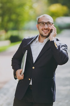 Homme d'affaires intelligent réussi en chemise blanche, costume classique, lunettes. l'homme se tient debout avec un ordinateur portable, parle sur un téléphone portable dans le parc de la ville à l'extérieur sur fond de nature. bureau mobile, concept d'entreprise.
