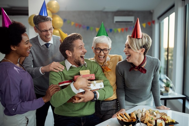 Homme d'affaires joyeux tenant un tas de cadeaux d'anniversaire et s'amusant avec des collègues lors d'une fête de bureau