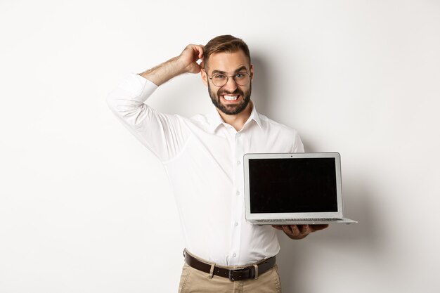 Homme d'affaires maladroit montrant l'écran de l'ordinateur portable et à la douteuse, debout contre un fond blanc inconfortable.