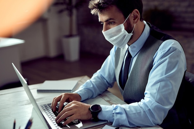 Homme d'affaires avec masque facial travaillant sur un ordinateur portable au bureau pendant la pandémie de coronavirus