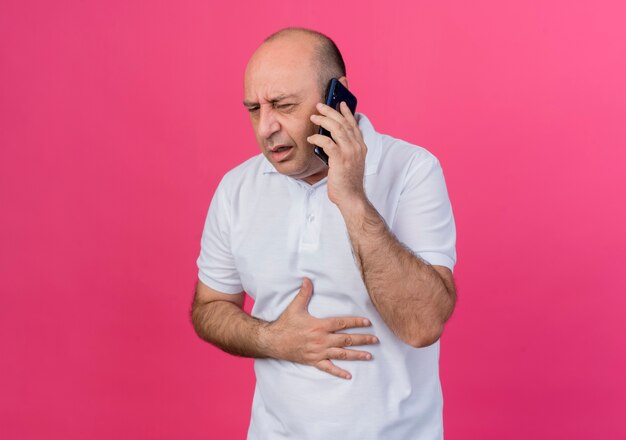 Homme d'affaires mature occasionnel douloureux, parler au téléphone à la recherche de mettre la main droite sur le ventre isolé sur fond rose avec espace de copie