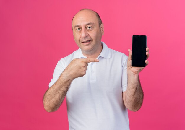 Homme d'affaires mature occasionnel impressionné montrant un téléphone mobile et pointant vers lui isolé sur fond rose