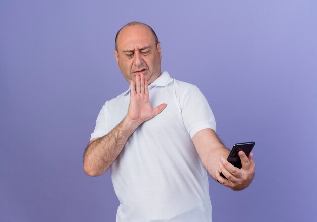 Homme d'affaires mature occasionnel non satisfait tenant et regardant le téléphone mobile et faisant des gestes non avec la main isolée sur fond violet avec espace copie