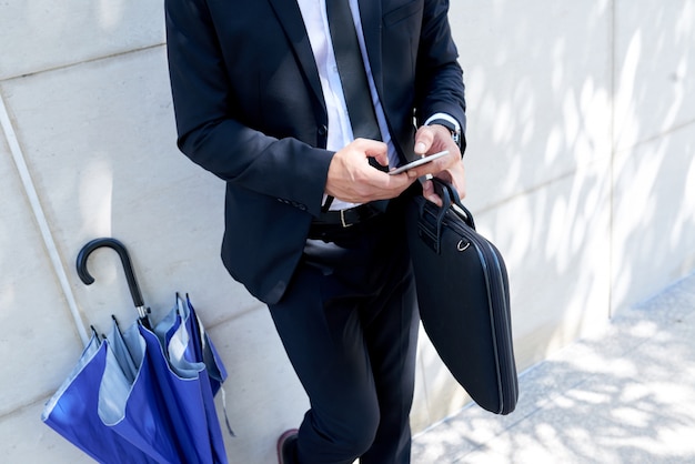 Homme d'affaires méconnaissable avec mallette et parapluie debout à l'extérieur et à l'aide de smartphone