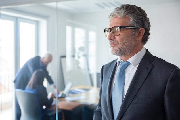 Homme d'affaires mûr pensif en costume formel et lunettes debout près du mur de verre du bureau, à la recherche de suite Copiez l'espace. Concept de portrait d'entreprise