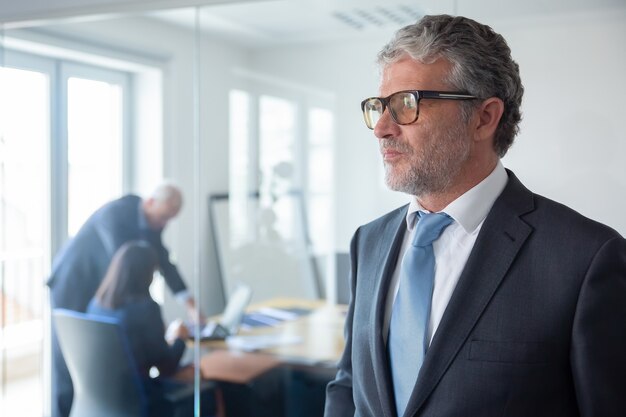 Homme d'affaires mûr pensif en costume formel et lunettes debout près du mur de verre du bureau, à la recherche de suite Copiez l'espace. Concept de portrait d'entreprise