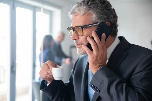 Homme d'affaires mûr pensif en sirotant un expresso dans une petite tasse en se tenant debout près du mur de verre du bureau et en parlant au téléphone mobile Copiez l'espace. Concept d'affaires et de pause-café